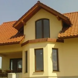 Jasnożółty dom jednorodzinny z brązową dachówką, drewnianymi elementami wykończeniowymi i trzema pionowymi oknami w wykuszu.